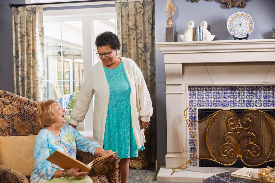 Two women in a cozy living room; one elderly woman sitting in a patterned armchair holding a book and smiling, while a standing woman in a turquoise dress and white cardigan looks at her warmly. Behind them is a fireplace with blue and white tiles and decorative items on the mantel.