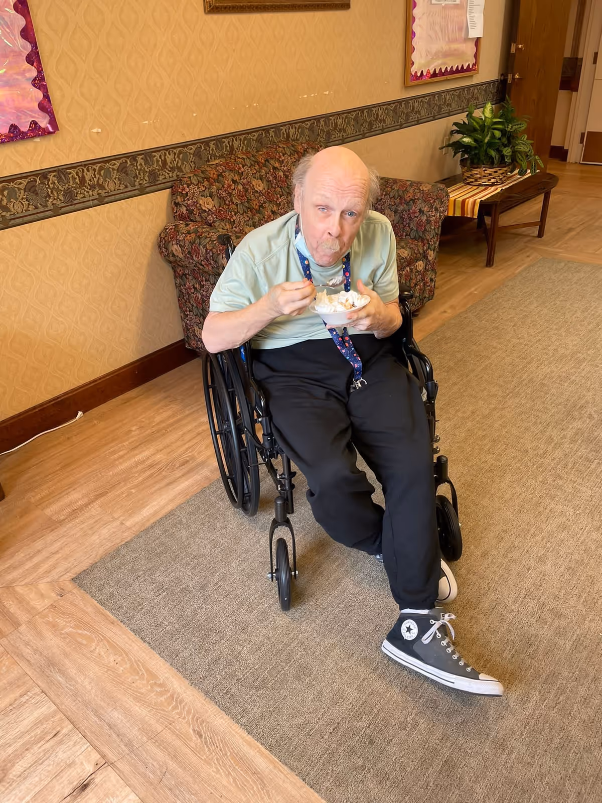 An elderly man in a wheelchair eating from a bowl with a spoon in a hallway of an assisted living facility. The hallway has wooden flooring, a beige patterned wallpaper with a decorative border, a floral-patterned couch, a wooden bench with a plant on it, and bulletin boards on the walls.