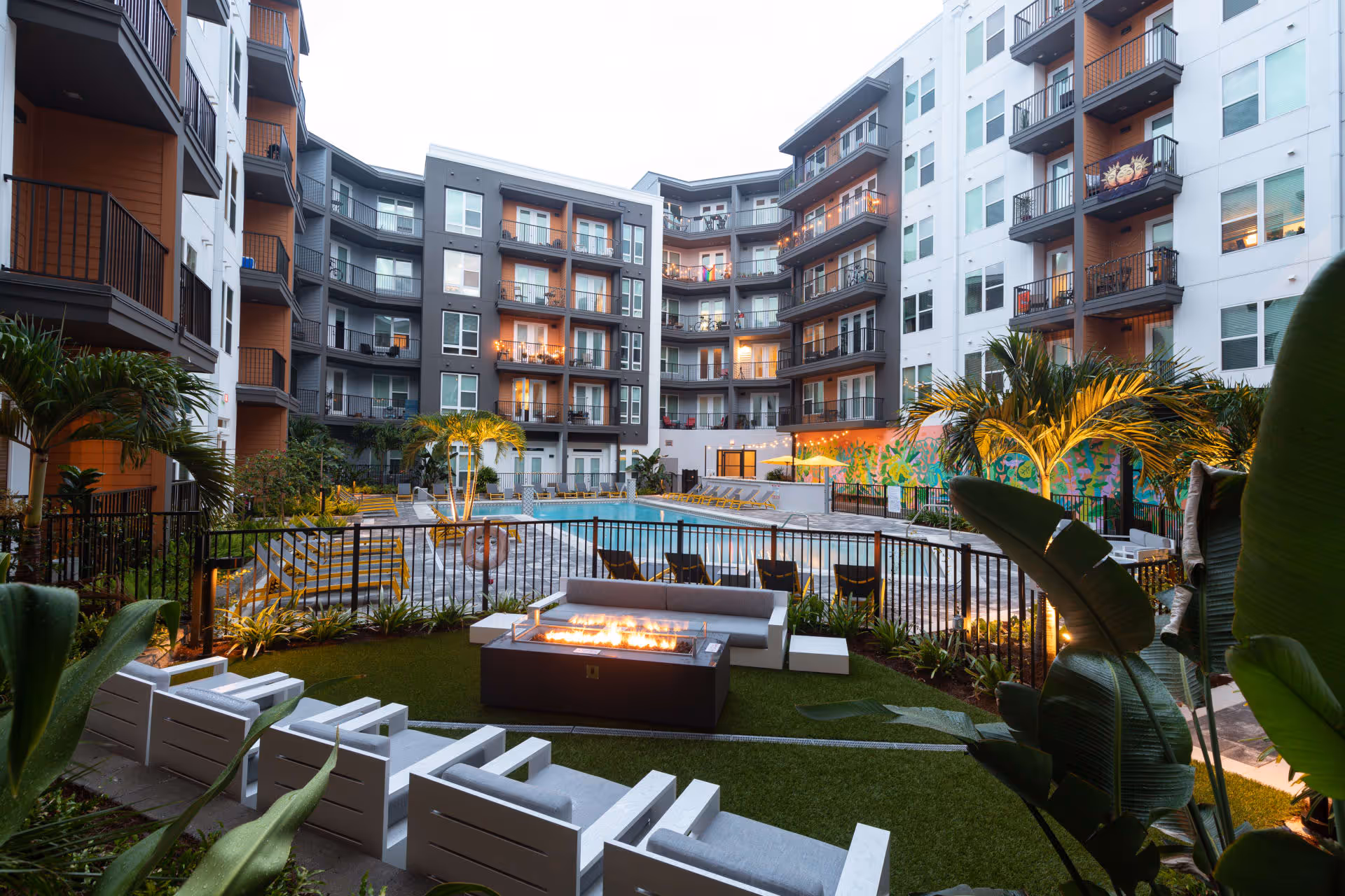 Outdoor courtyard area of a multi-story residential building with balconies. The courtyard features a swimming pool surrounded by lounge chairs, palm trees, and a seating area with cushioned chairs and a fire pit. The building has a modern design with gray and white exterior walls accented by orange panels.