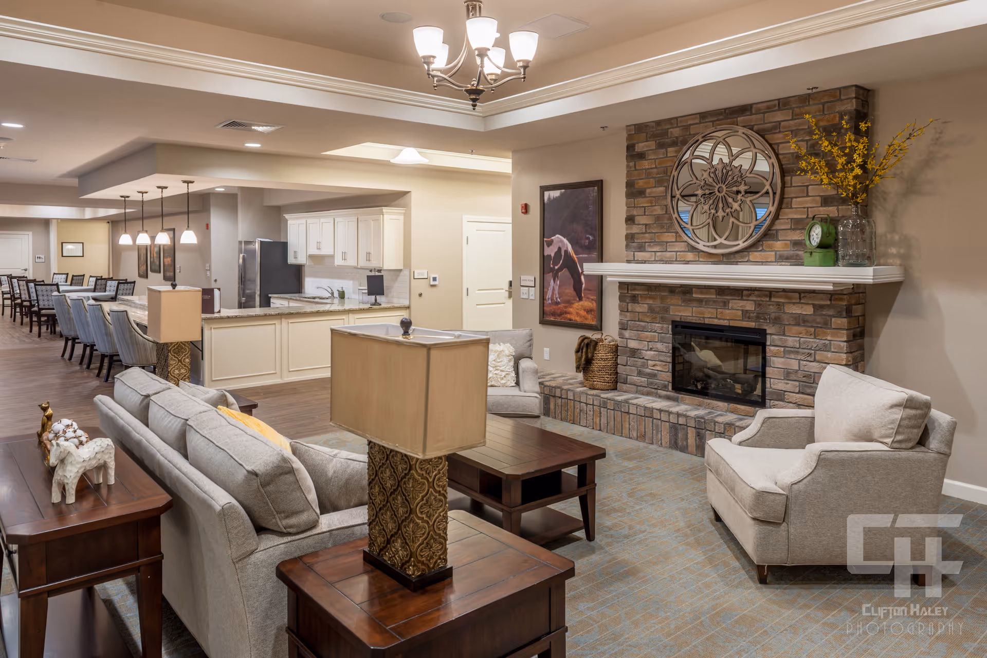 A cozy senior living common area featuring a seating arrangement with a sofa, armchairs, and wooden tables with lamps. A brick fireplace with a decorative mirror and a vase with yellow flowers is on the right wall. In the background, there is a kitchen area with white cabinets and a dining area with multiple tables and chairs. The space is warmly lit with ceiling lights and pendant lamps.