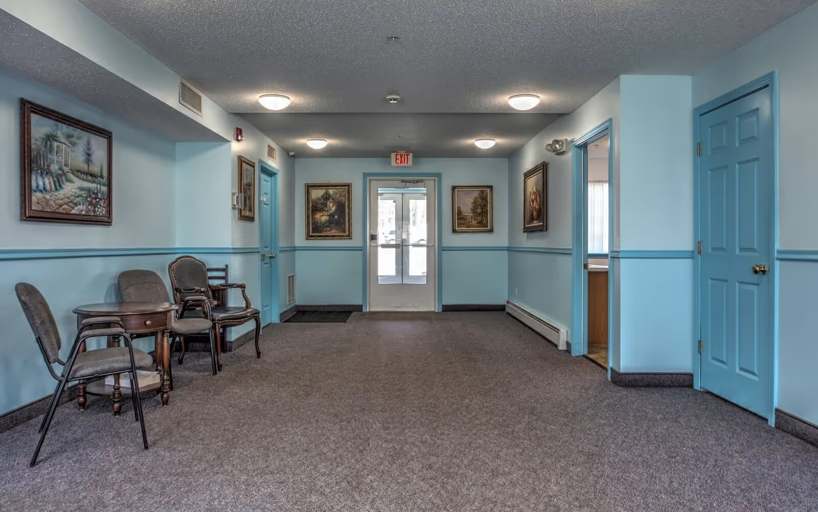 A hallway in a senior living facility with light blue walls and doors, carpeted floor, several chairs and a small wooden table on the left side, framed paintings on the walls, and a glass exit door at the end of the hallway.