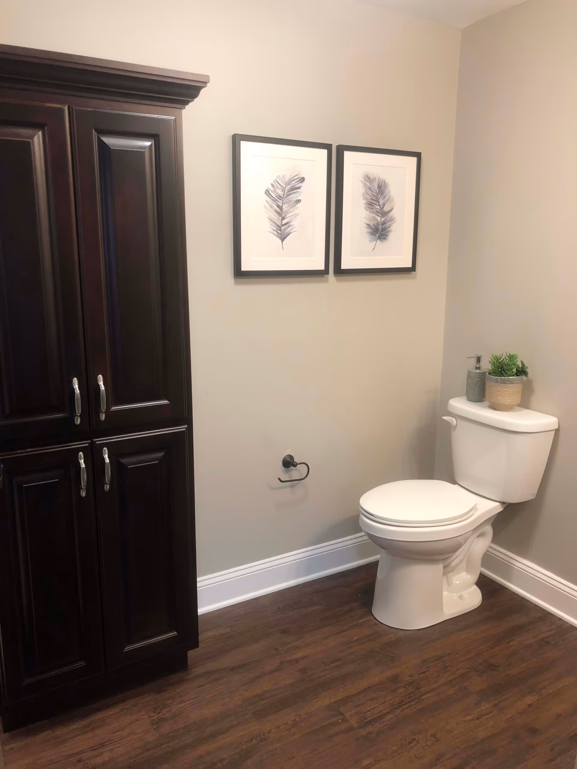 A bathroom corner featuring a white toilet with a soap dispenser and a small potted plant on the tank. To the left is a tall dark wooden cabinet with silver handles. Above the toilet on the beige wall are two framed pictures of feathers. The floor is a dark wood finish.