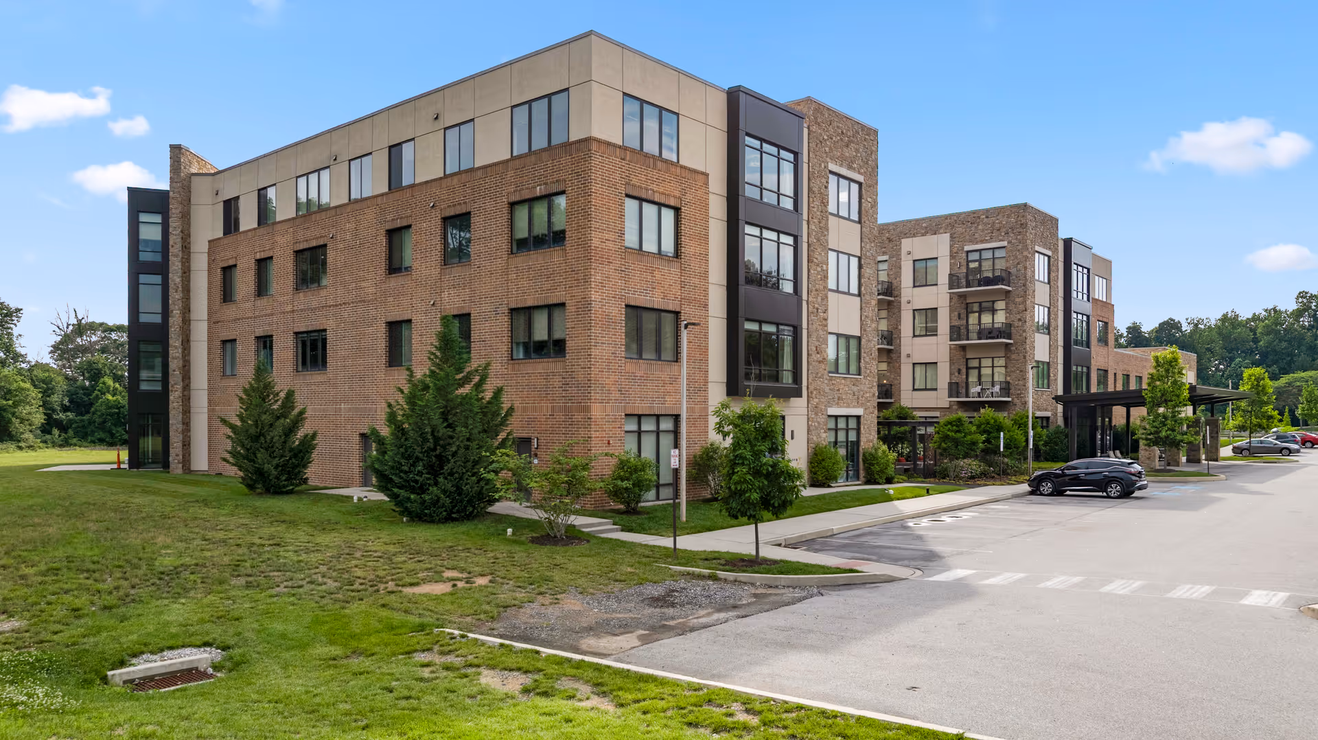 Exterior view of a multi-story brick senior living building with parking, trees, and a driveway under a blue sky.