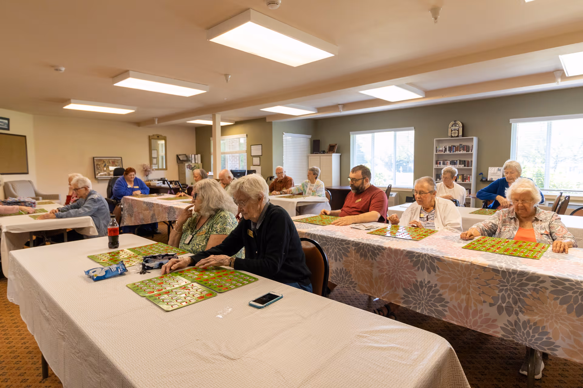 A group of elderly people sitting at tables in a well-lit room playing bingo. The tables are covered with tablecloths, and each person has bingo cards in front of them. The room has large windows, bookshelves, and overhead lighting.