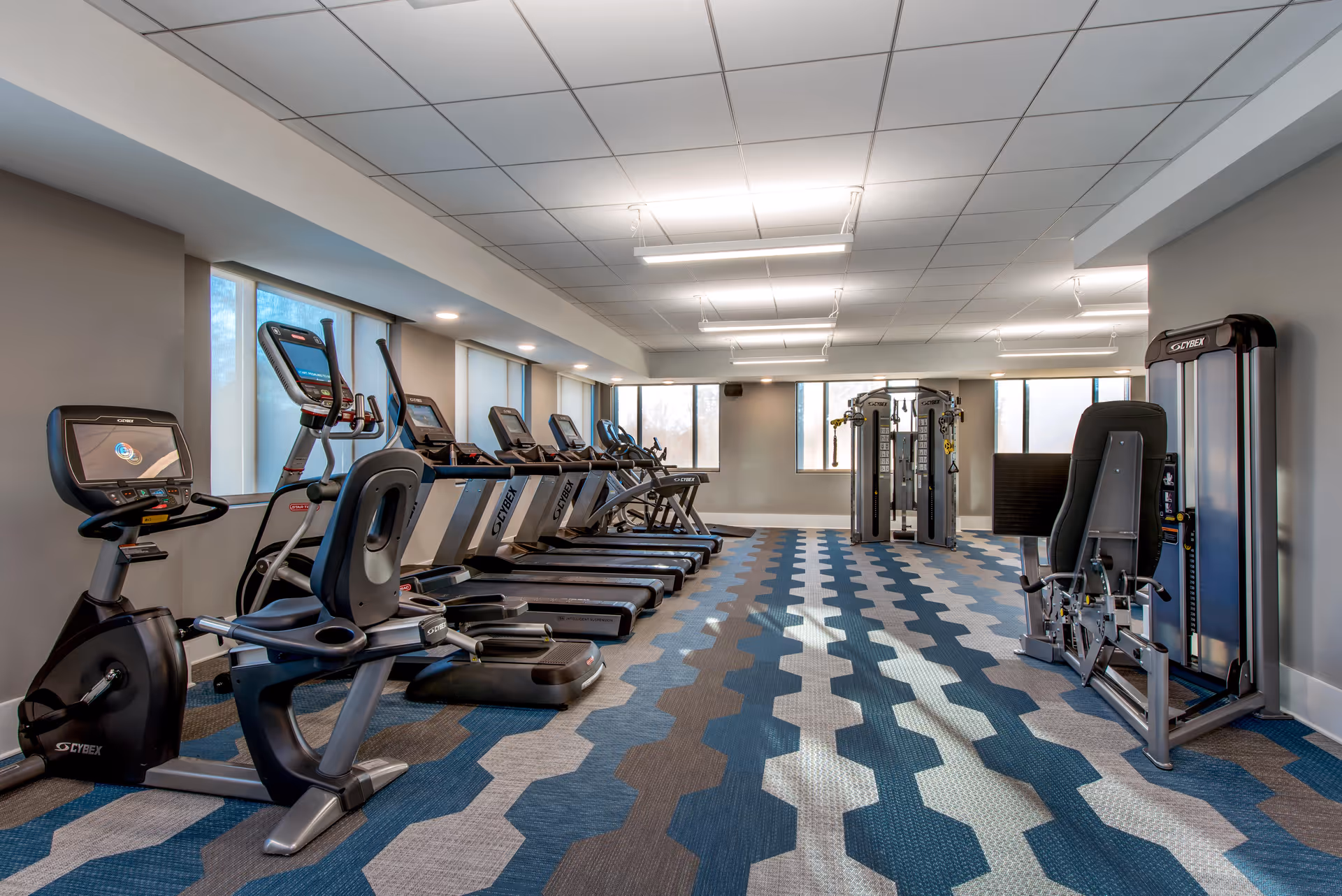 Well-lit fitness room with treadmills, stationary bikes, and weight machines on patterned carpeting.