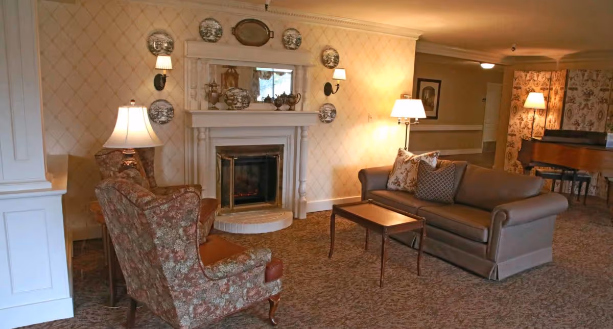 A cozy living room area in Delaware Plaza Assisted Living Community featuring a beige sofa with patterned cushions, two floral upholstered armchairs, a wooden coffee table, and a white fireplace adorned with decorative plates and silver tea sets. The room is softly lit by several lamps and has patterned wallpaper and carpet.