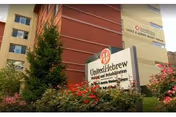 Exterior view of the United Hebrew of New Rochelle building with a sign in front surrounded by flowers and greenery.