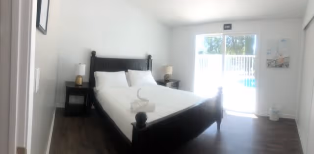 A bright bedroom with a dark wooden bed frame and white bedding. Two white towels are folded on the bed. There are two bedside tables with lamps on either side of the bed. A large window or glass door lets in natural light and shows a glimpse of an outdoor pool area. The floor is dark wood, and the walls are white with minimal decoration.