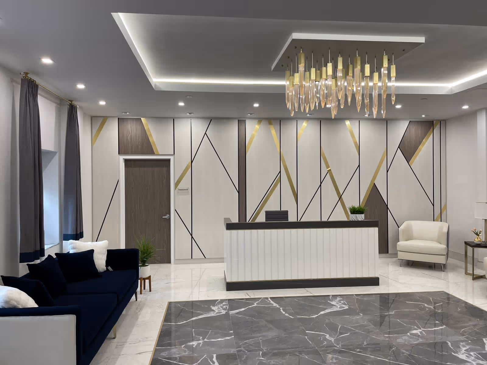 Bright modern reception lobby with a front desk, a chandelier, navy sofa and armchair in front of a geometric accent wall.