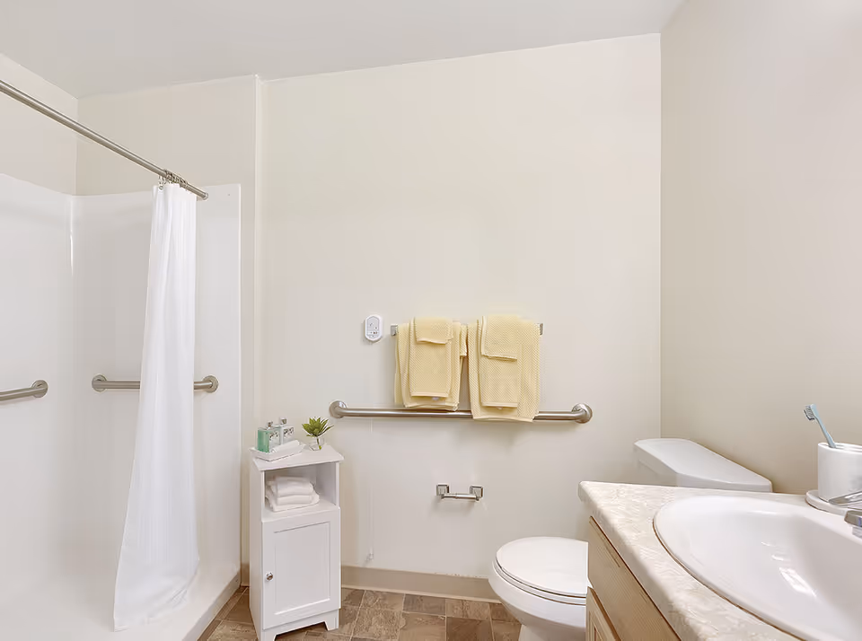 A clean and simple bathroom with a white shower curtain, grab bars on the walls, a white toilet, a sink with a countertop, and a small white cabinet holding towels and toiletries. Two yellow towels hang on a grab bar above the toilet paper holder.