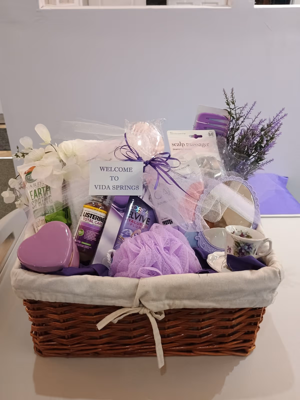 Wicker gift basket on a table filled with purple-themed toiletries, flowers, and a 'WELCOME TO VIDA SPRINGS' card.