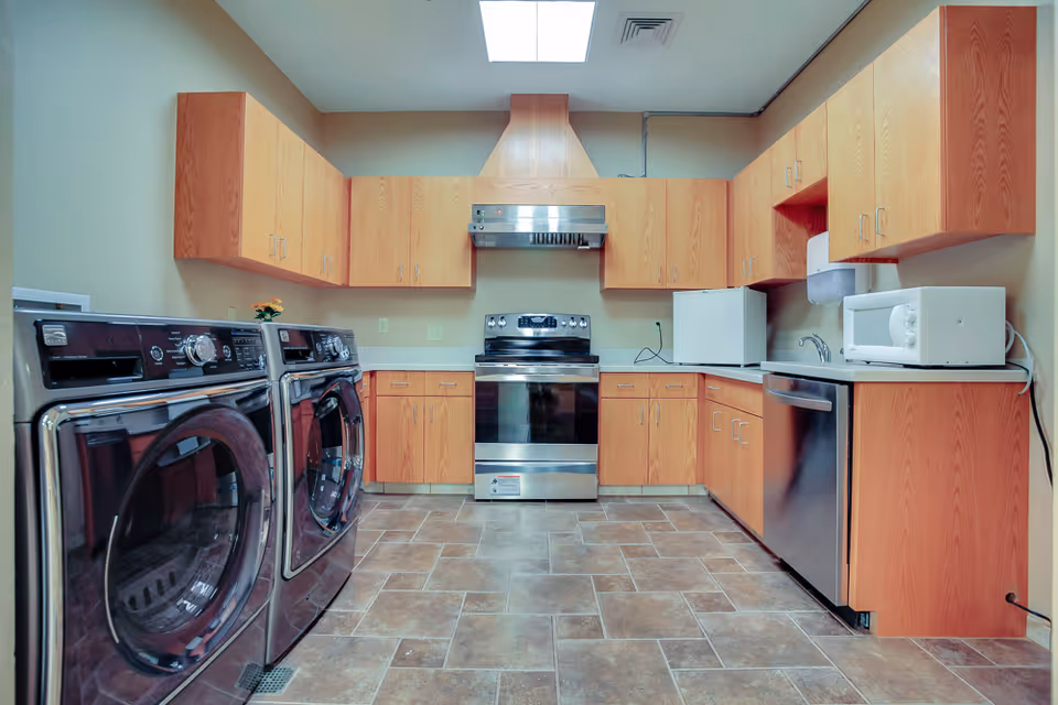 A spacious kitchen area with wooden cabinets, a stainless steel stove with an overhead vent, a dishwasher, a microwave, a small refrigerator, and a washer and dryer set. The floor is tiled and there is a skylight on the ceiling providing natural light.