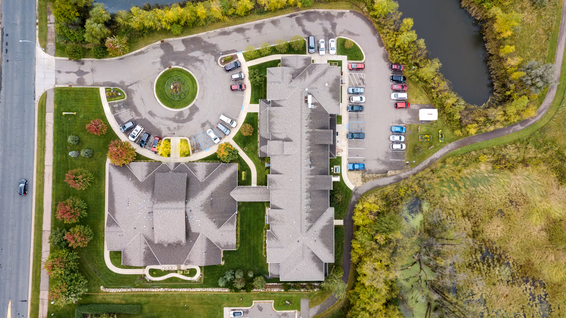 Aerial view of Legacy of Delano Senior Living facility showing the building rooftops, surrounding parking lots with cars, circular driveway, landscaped green areas with trees, and a nearby pond with walking paths.