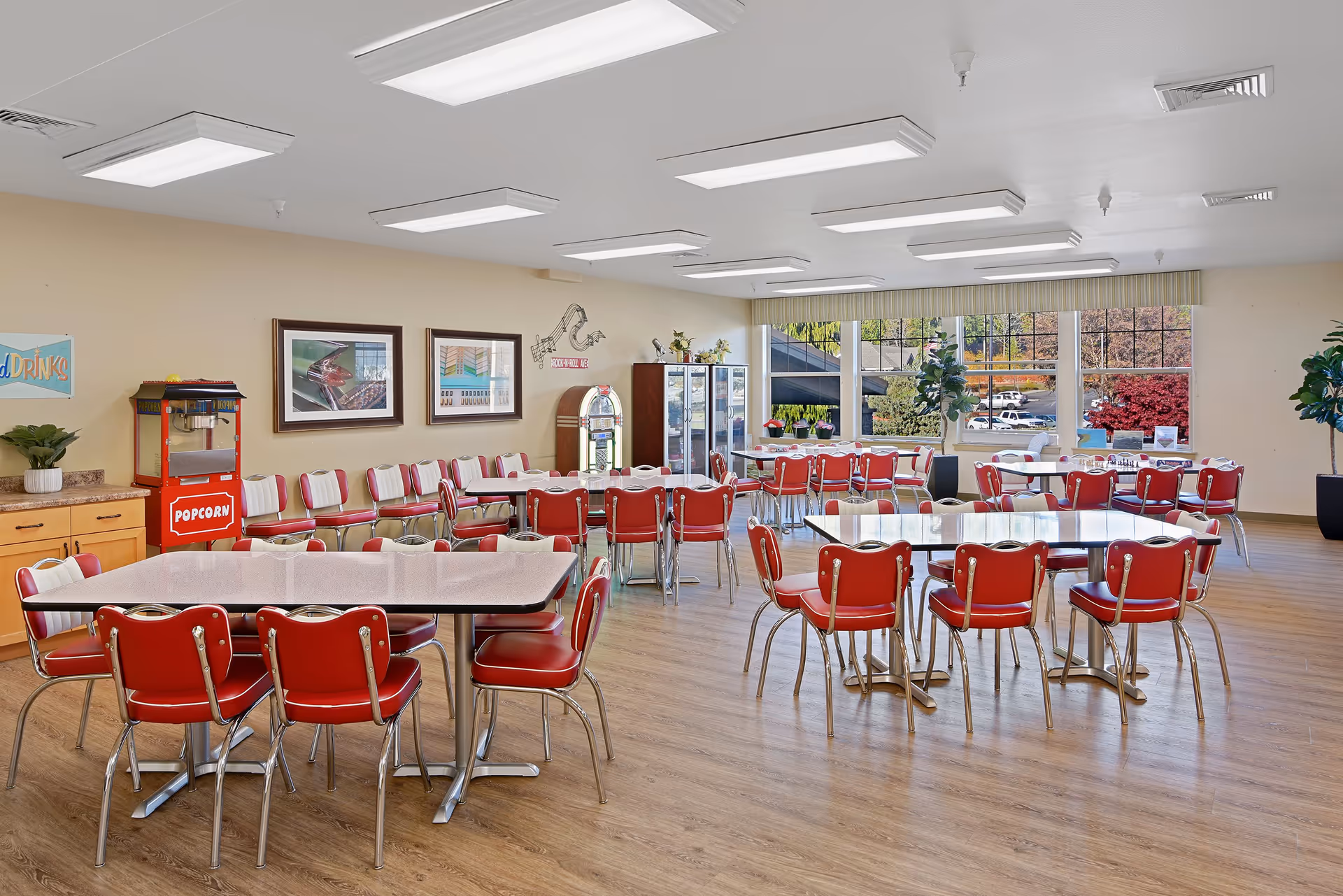 A spacious dining room with multiple tables and red and white retro-style chairs. The room features large windows letting in natural light, a popcorn machine, a jukebox, framed pictures on the wall, and plants near the windows. The floor is wooden and the ceiling has several rectangular fluorescent lights.