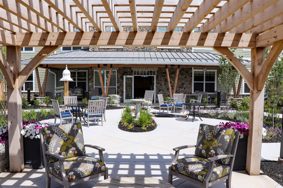 Outdoor seating area at a senior living facility with wooden pergola overhead, cushioned chairs with floral patterns, round tables, and a birdhouse. The background shows a building with stone and siding exterior, windows, and a door leading inside. There are landscaped plants and flowers around the paved patio area.
