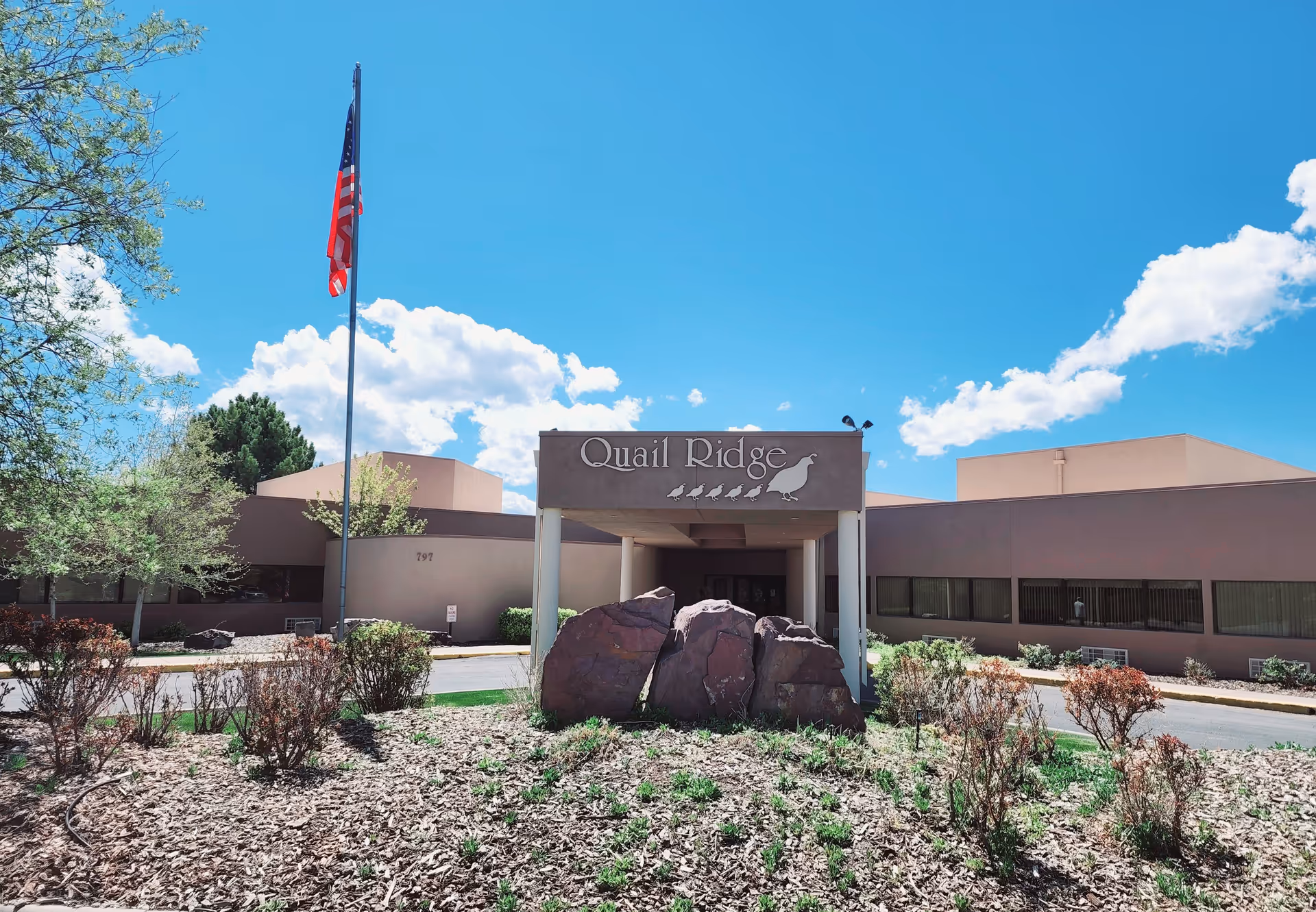 Front exterior view of Quail Ridge facility with a covered entrance, large rocks in the landscaped area, an American flag on a flagpole, and a clear blue sky with some clouds.