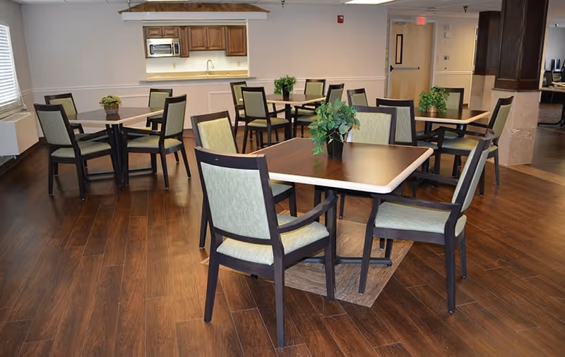 Communal dining room with several square tables, green-upholstered chairs, potted plants, and a serving counter with a microwave in the background.