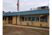 Front view of a one-story beige building with a teal metal roof, large windows, a sidewalk, and an American flag on a flagpole in front.