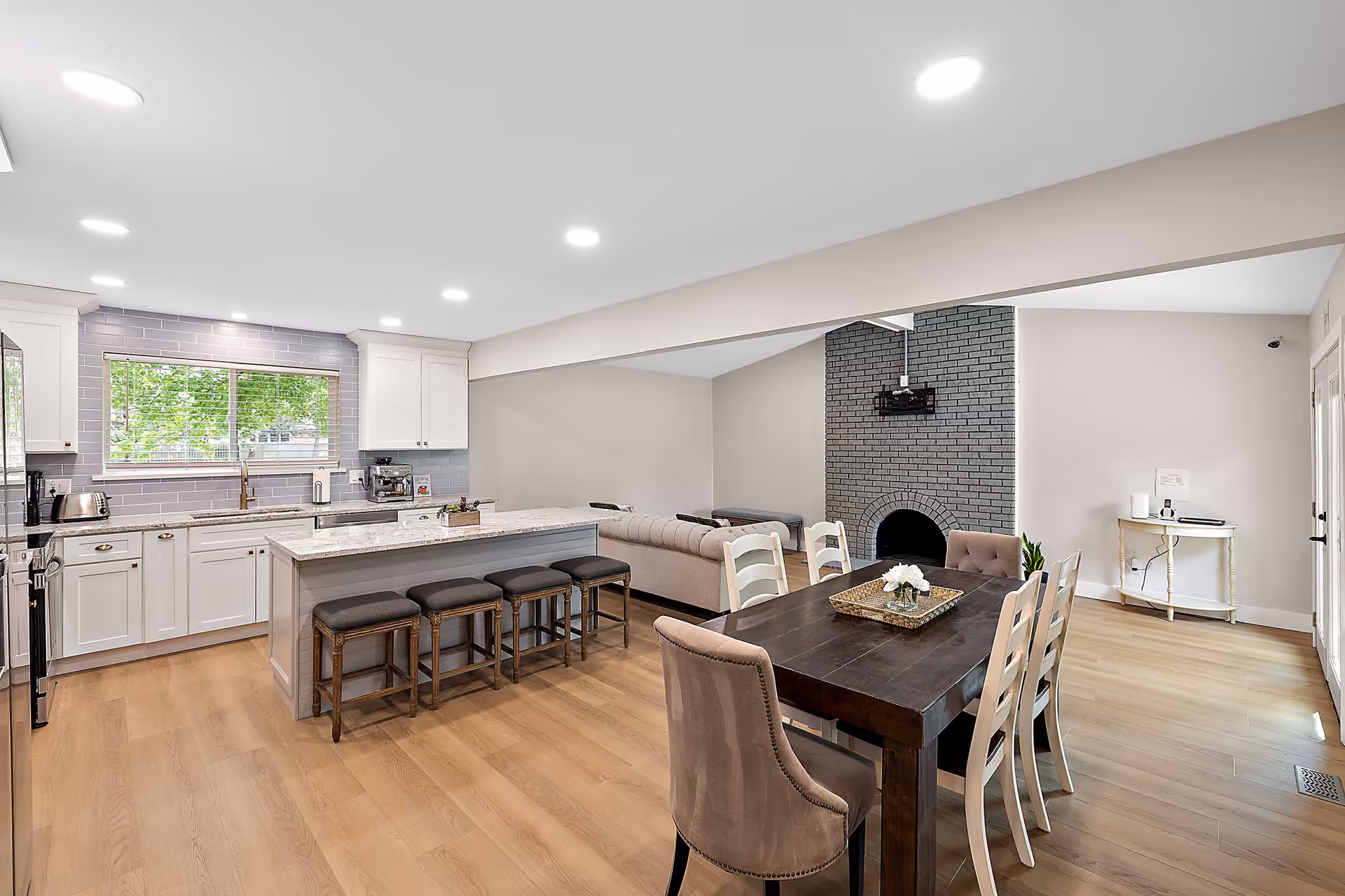 Open concept interior space featuring a modern kitchen with white cabinets, a large island with four stools, and a window with a view of green trees. Adjacent to the kitchen is a dining area with a dark wooden table and six chairs, and a living area with a beige sofa and a gray brick fireplace with a mounted TV bracket above it. The room has light wood flooring and recessed ceiling lights.