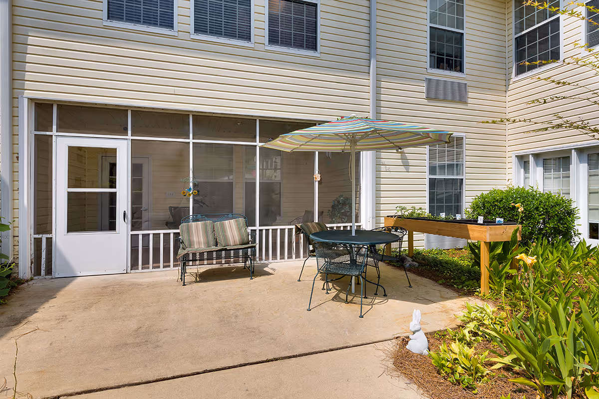 Outdoor patio area at Lakeview Estates - Memory Care Community featuring a metal table with four chairs and a striped umbrella, a metal bench with striped cushions, a raised wooden planter box with plants, and surrounding greenery including bushes and flowers. The patio is adjacent to a beige building with multiple windows and a screened porch.