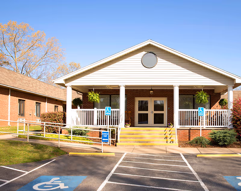 Front exterior of a brick senior living center with a covered entrance, ramp and steps, and handicap parking signs.