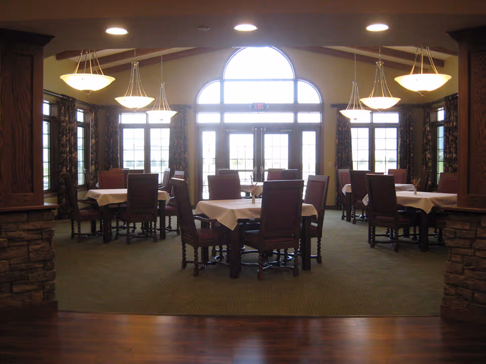 A dining room with multiple tables covered with white tablecloths and surrounded by wooden chairs with red cushions. The room features large windows with floral curtains and an arched window above double glass doors letting in natural light. Several pendant lights hang from the ceiling, and the floor is carpeted with a green rug, bordered by wooden flooring.