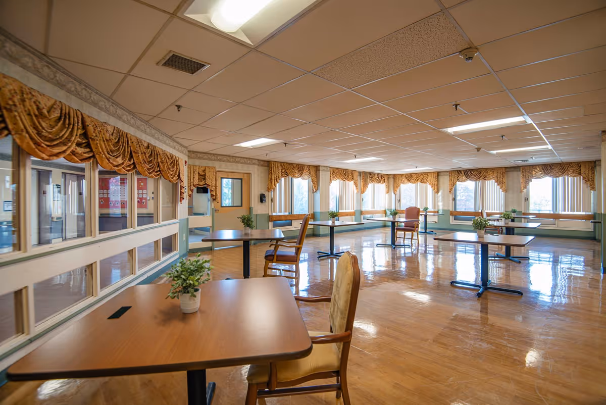 A spacious, well-lit dining room with multiple wooden tables and chairs arranged neatly. The room features large windows with gold-colored drapes, allowing natural light to fill the space. Small potted plants are placed on the tables, and the floor has a polished wooden finish.