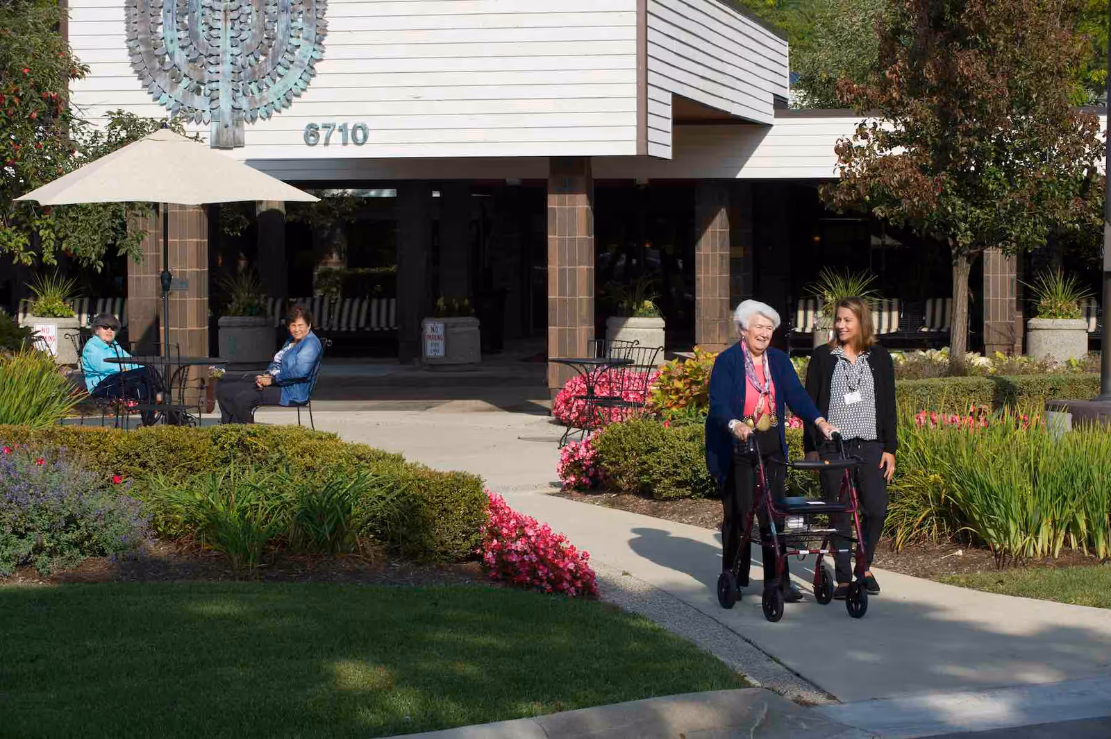An elderly woman using a walker is accompanied by a younger woman walking on a sidewalk outside a building with the number 6710. Two other women are seated at a table under an umbrella near the entrance. The area is landscaped with green bushes, pink flowers, and trees.