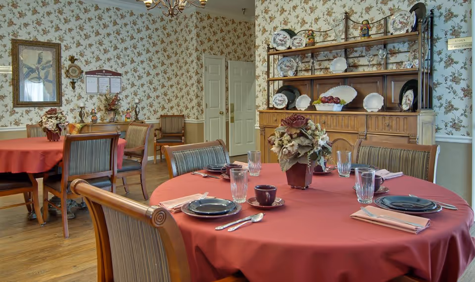 A dining room with round tables covered in red tablecloths, set with plates, cups, glasses, and silverware. There are wooden chairs with striped cushions around the tables. The room has floral wallpaper and wooden flooring. A wooden hutch displaying decorative plates and a floral centerpiece on the table are also visible.