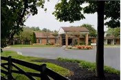 Exterior view of a single-story brick building with a covered entrance, surrounded by trees and landscaping, with a paved driveway and a wooden fence in the foreground.