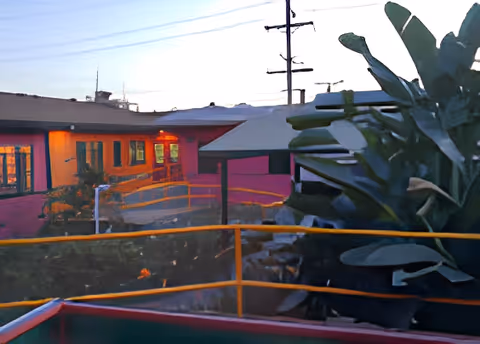Outdoor view of a nursing care center building at dusk with illuminated windows, a ramp with yellow railings, and large green plants in the foreground.
