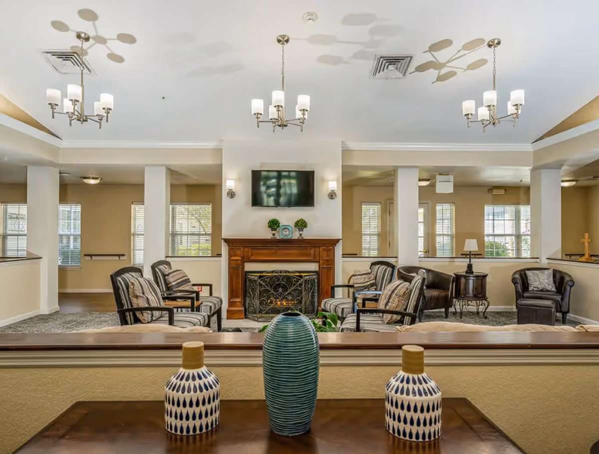 A cozy living room area with a fireplace and a mounted flat-screen TV above it. The room features multiple armchairs with striped cushions arranged around the fireplace. There are three chandeliers hanging from the ceiling, and several windows letting in natural light. In the foreground, there is a wooden table with three decorative vases.