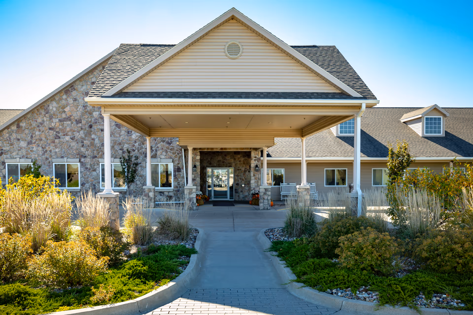 Front entrance of a single-story assisted living building with a covered porte-cochere, stone and siding exterior and landscaped walkway.