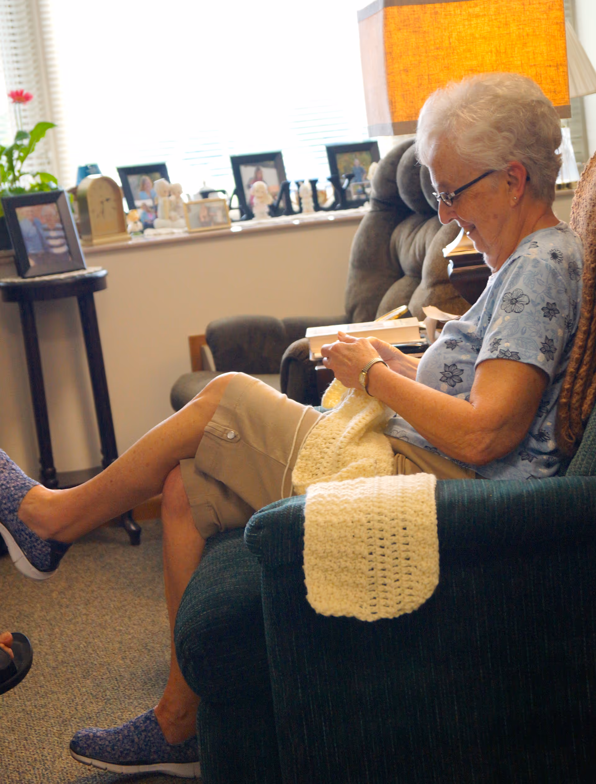 An elderly woman with short white hair and glasses is sitting comfortably in a green armchair, knitting a cream-colored blanket. She is smiling and appears relaxed. The room has a cozy atmosphere with framed photos and decorative items on a windowsill behind her, a side table with a lamp, and another armchair in the background.