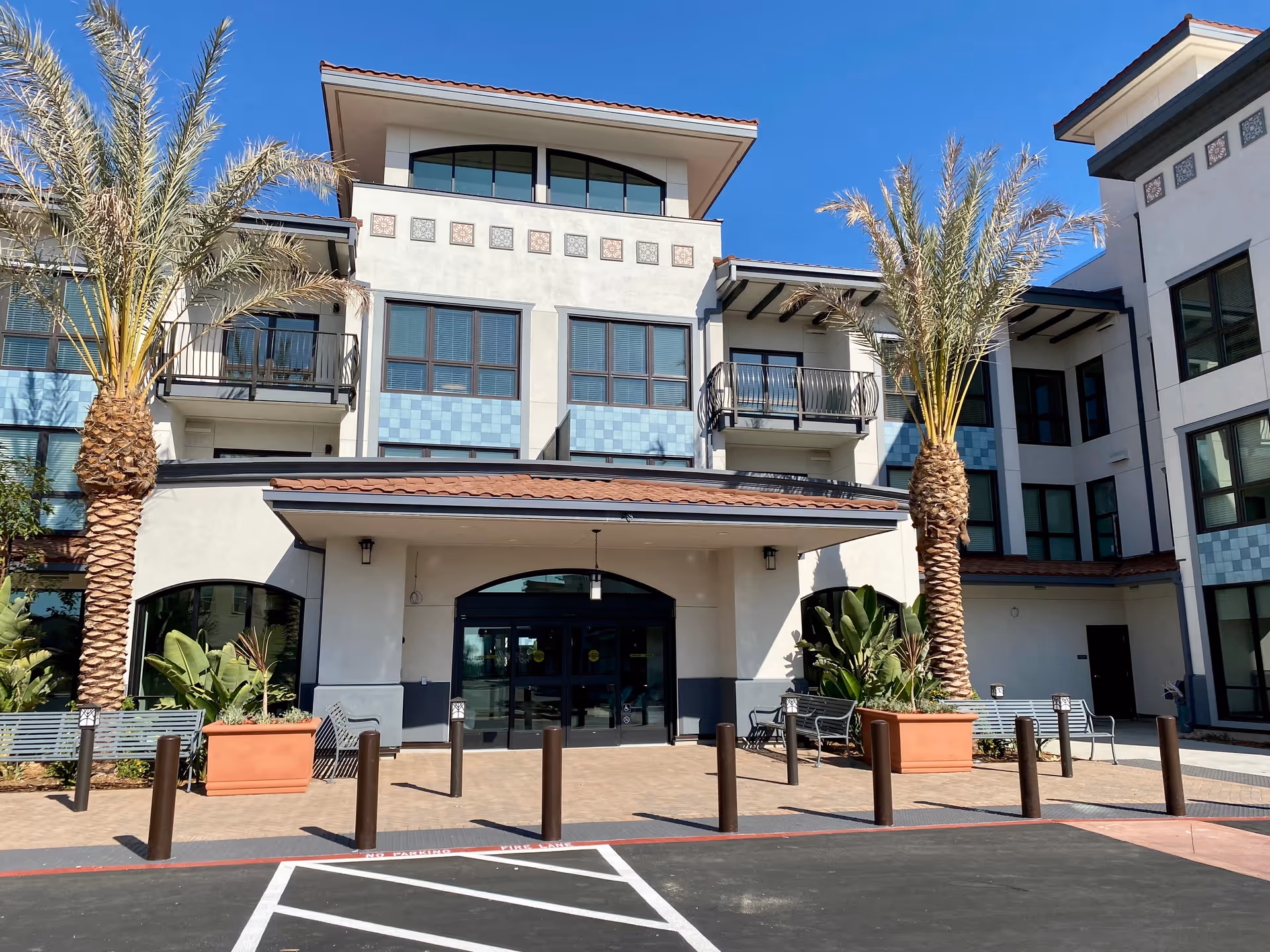 Exterior front view of Westmont of Cypress senior living facility with a clear blue sky, palm trees, benches, and a covered entrance with glass doors.