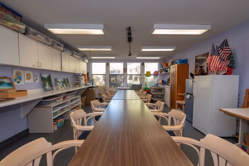 A well-lit activity room with a long wooden table surrounded by beige chairs. The room has large windows at the far end letting in natural light. On the left wall, there are white cabinets and shelves holding various art supplies and paintings. On the right side, there are storage units, a refrigerator, and several American flags displayed. The walls are painted light purple.