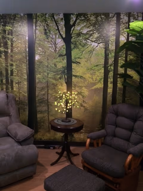 Cozy seating area with two armchairs, a small round table holding a lit decorative tree, and a forest mural on the wall.