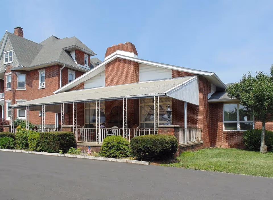 Exterior view of a brick building with a covered porch and decorative metal railings. The building has a sloped roof with a chimney and is surrounded by trimmed bushes and a small tree. The sky is clear and blue.