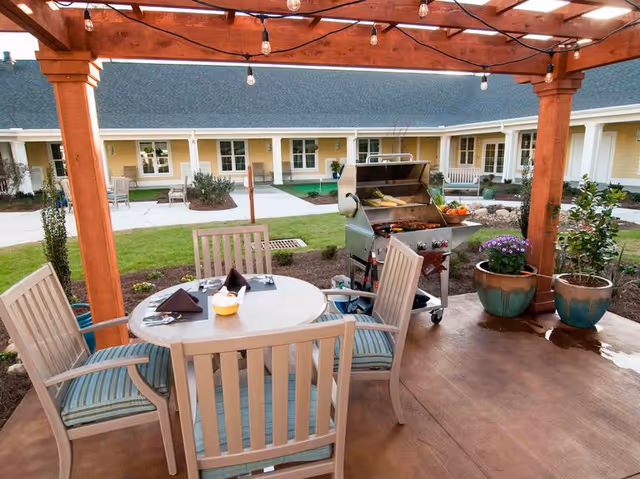 Outdoor patio area with a wooden pergola overhead, string lights hanging from the pergola, a round table with four chairs around it, and a grill with food cooking. There are potted plants around the patio and a courtyard with grass and walkways in the background, along with a yellow building with white trim and multiple windows and doors.