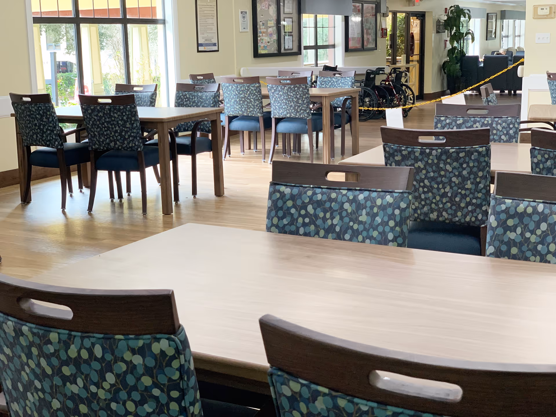 A bright dining area with multiple wooden tables and chairs featuring blue patterned upholstery. Large windows allow natural light to fill the room. In the background, there are framed pictures on the walls, a wheelchair, and a yellow chain blocking access to a hallway.
