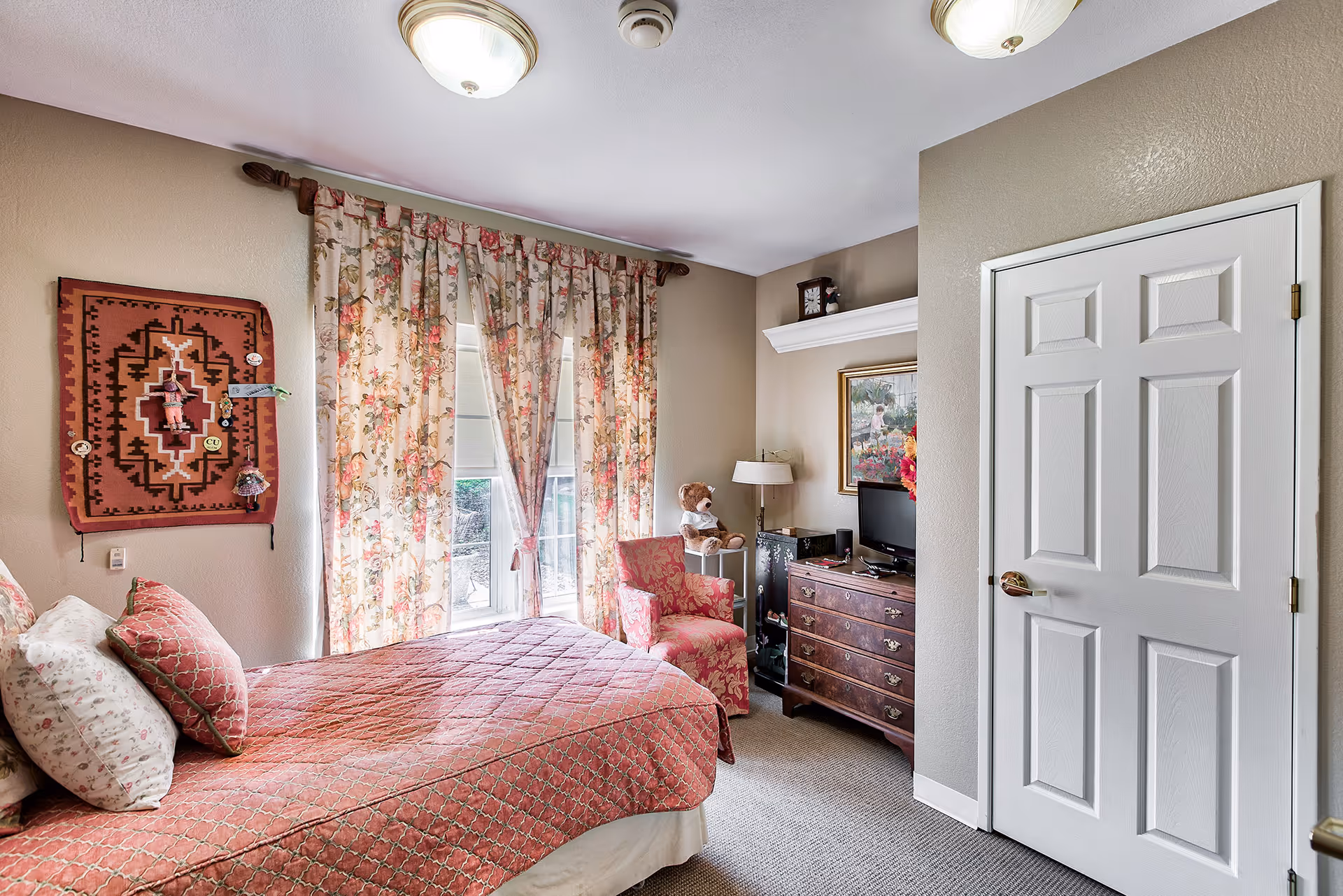 A cozy bedroom with a bed covered in a red patterned quilt and floral pillows. There is a window with floral curtains letting in natural light. A red upholstered armchair sits next to a small table with a teddy bear on it. A wooden dresser with a small TV and a lamp is against the wall, along with a framed painting above it. The walls are painted beige and there is a white door on the right side.