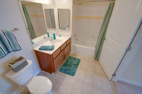 A clean bathroom with a white toilet, a wooden vanity with a white countertop, a large mirror above the sink, a green bath mat on the tiled floor, a bathtub with a green shower curtain, and a striped towel hanging on a rack.