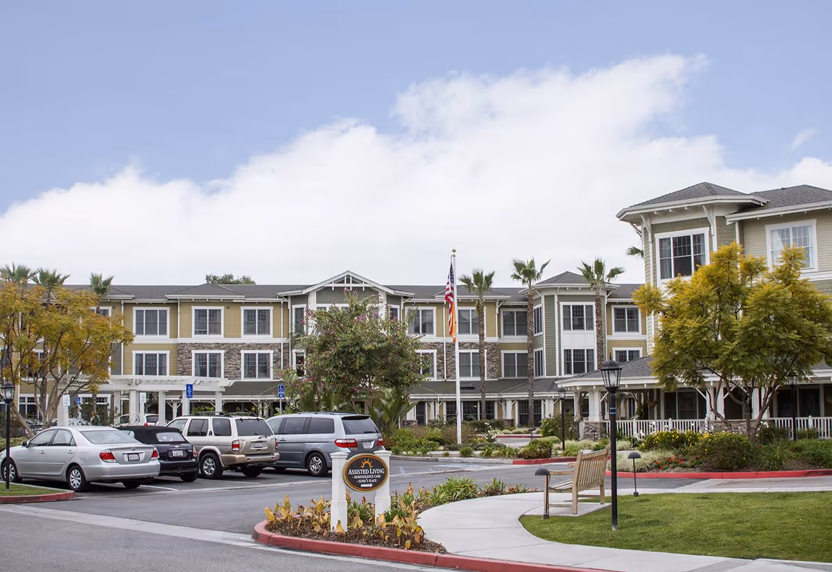 Exterior view of Ivy Park at Seal Beach, a multi-story assisted living facility with beige and stone facade, surrounded by palm trees and landscaped greenery. Several parked cars are visible in the parking lot in front of the building, along with a curved sidewalk and a bench near a lamp post.