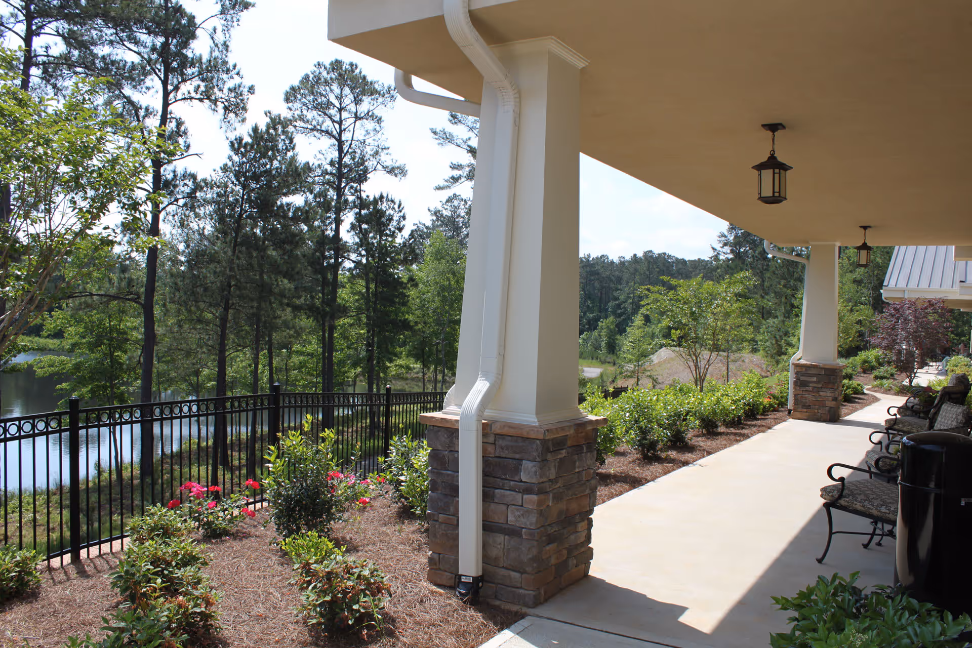 Covered outdoor patio area with stone pillars and hanging lanterns, overlooking a landscaped garden with bushes and flowers, black metal fence, and a pond surrounded by tall pine trees in the background.
