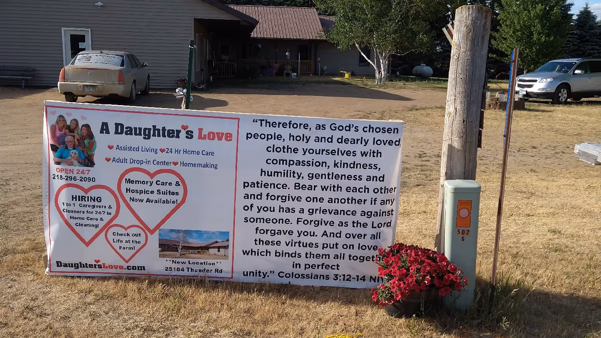 Outdoor scene showing a large sign for 'A Daughter's Love' facility placed on a grassy area near a wooden post with a flower pot of red flowers. The sign advertises assisted living, 24-hour home care, adult drop-in center, homemaking, memory care, hospice suites, and hiring caregivers and cleaners. A house and parked cars are visible in the background.