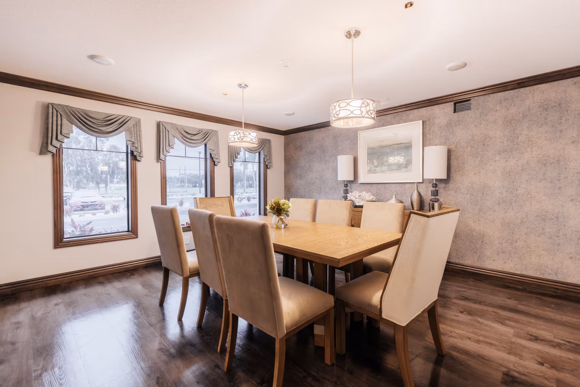 A dining room with a wooden table surrounded by eight beige upholstered chairs. The room has three large windows with decorative valances, two pendant lights hanging above the table, and a sideboard against a textured wall with two lamps and framed artwork. The floor is dark wood.