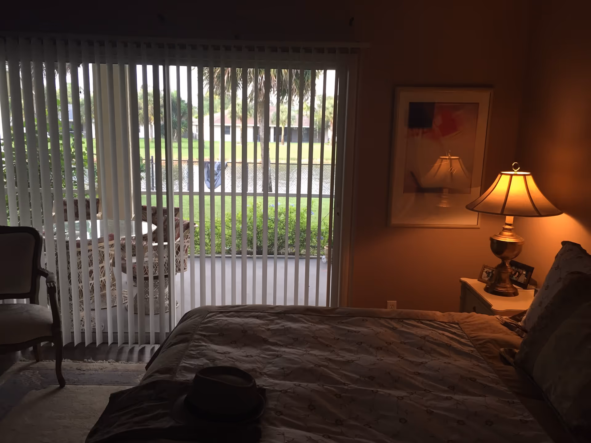 Bedroom with a bed in the foreground, a lit bedside lamp, and vertical blinds covering sliding glass doors that open to a patio and pond view.