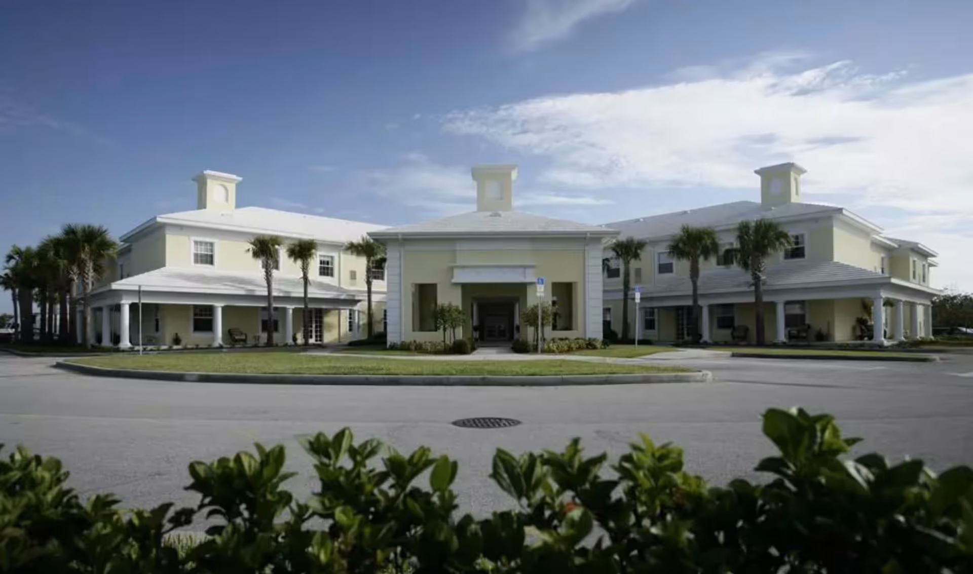 Exterior view of a two-story assisted living facility building with a light yellow facade, white roof, and multiple palm trees in front. The building has a covered entrance and a circular driveway with greenery in the foreground under a partly cloudy sky.