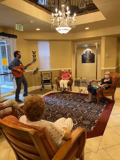 A small group of elderly people seated in a cozy common area with cushioned chairs arranged around a patterned rug. A man wearing a mask is standing and playing a guitar near the entrance door. The room has warm lighting with a chandelier hanging from the ceiling and a decorative water feature on one wall.