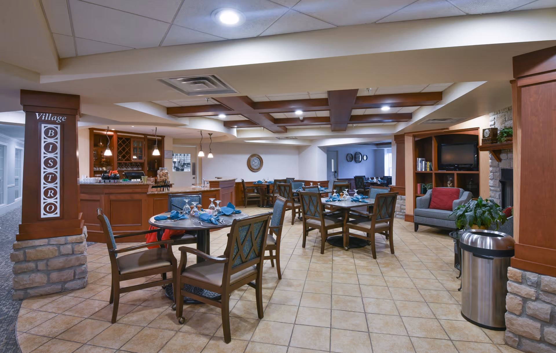 Interior view of a dining area named Village Bistro with several round tables set with blue napkins, glasses, and silverware. The room features wooden furniture, a small bar area with hanging lights, a cozy seating area with a TV and fireplace, and tiled flooring.
